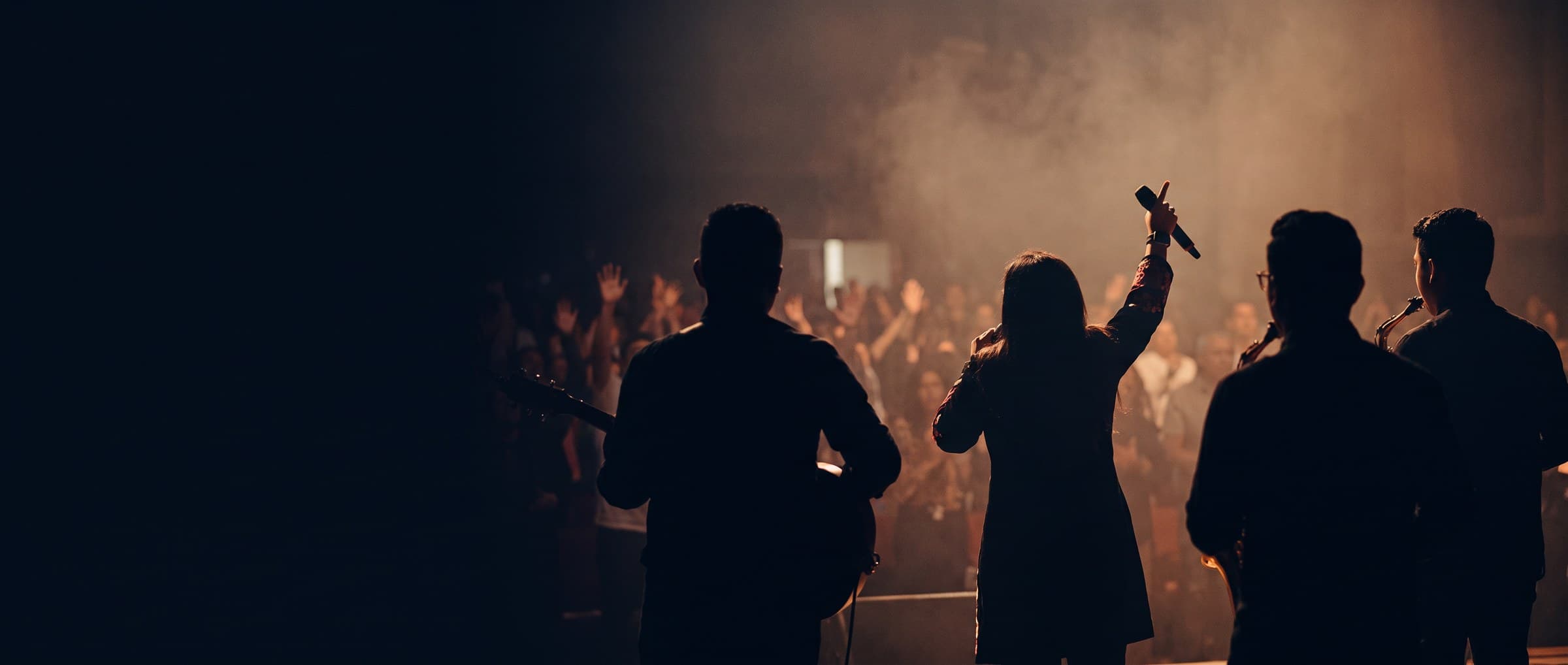 Worship leader with raised hand and band on stage, facing a crowd under warm light